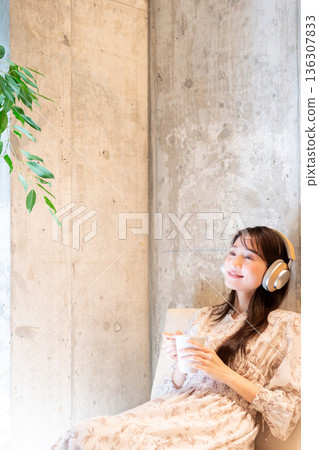 一位年輕女子放鬆地躺在沙發上,戴著耳機聽音樂,喝咖啡。 一位年輕女子放鬆地躺在沙發上,戴著耳機聽音樂,喝咖啡。 136307833