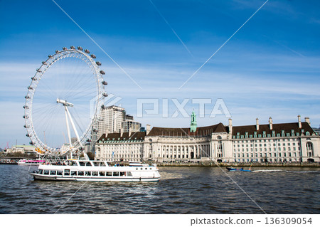 The London Eye, a large Ferris wheel built along the River Thames that flows through London under a blue sky with thin clouds 136309054