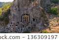 Aerial view of ancient Lycian rock tombs carved into mountain cliff in Fethiye, Turkey. Historic archaeological site surrounded by forest and rugged terrain. 136309197