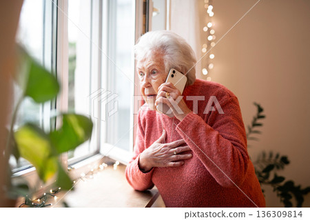 Old woman standing by window, making phone call. Old woman standing by window, making phone call. 136309814