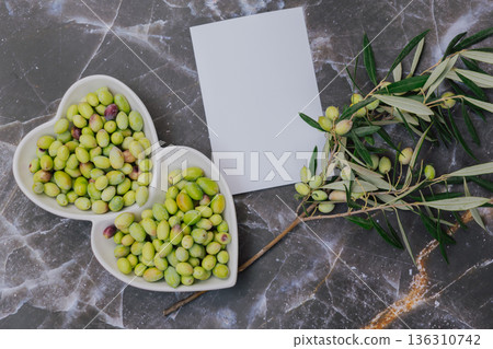 Freshly harvested green olives in heart shaped white plate with olive tree branch, blank paper for copy space, top view Mediterranean food concept Freshly harvested green olives in heart shaped white plate with olive tree branch, blank paper for copy space, top view Mediterranean food concept 136310742