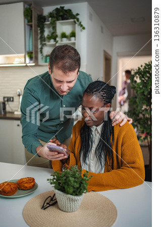 Diverse couple using smartphone at home kitchen table Diverse couple using smartphone at home kitchen table 136310879