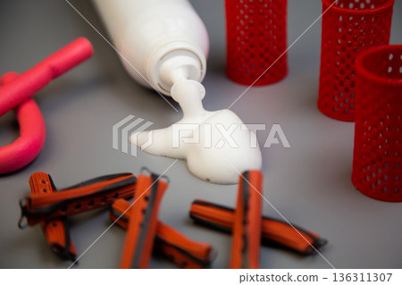A close-up on a gray background shows a bottle of foam for long-term hair styling, white foam poured out of it, next to it there are curlers of different shapes and sizes. The concept of styling 136311307