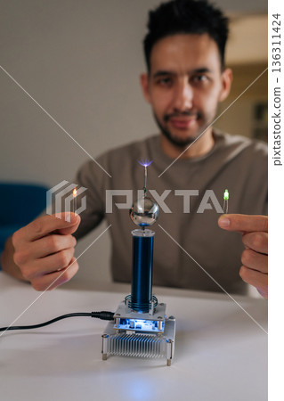 Vertical portrait of young man demonstrating tesla coil wireless power transmission, illuminating two light bulbs without direct contact, showcasing electricity, science and hobby experiment. Vertical portrait of young man demonstrating tesla coil wireless power transmission, illuminating two light bulbs without direct contact, showcasing electricity, science and hobby experiment. 136311424
