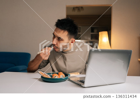 Bearded young man eating Asian delivery food with chopsticks while using laptop at home, combining dinner and work. Concept of modern multitasking lifestyle, domestic comfort and remote activity. 136311434