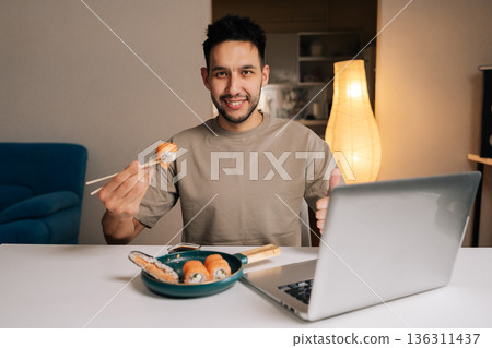 Friendly young man holding sushi with chopsticks, sitting at desk with laptop, smiling looking at camera and giving thumbs up gesture, indicating satisfaction working from home or enjoying meal. Friendly young man holding sushi with chopsticks, sitting at desk with laptop, smiling looking at camera and giving thumbs up gesture, indicating satisfaction working from home or enjoying meal. 136311437