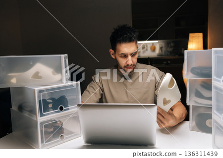 Portrait of male entrepreneur organizing shoe inventory at home, managing online store and e-commerce business using laptop, ensuring efficient storage, preparing for shipping. 136311439