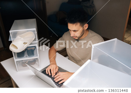 Top view of male entrepreneur managing online shoe store from home, organizing new footwear in clear storage boxes and using typing laptop for business operations, order fulfillment digital marketing. Top view of male entrepreneur managing online shoe store from home, organizing new footwear in clear storage boxes and using typing laptop for business operations, order fulfillment digital marketing. 136311448