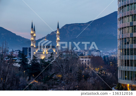 Illuminated mosque with minarets rising above city park and modern buildings at dusk with mountain backdrop and evening atmosphere, Tirana, Albania Illuminated mosque with minarets rising above city park and modern buildings at dusk with mountain backdrop and evening atmosphere, Tirana, Albania 136312016