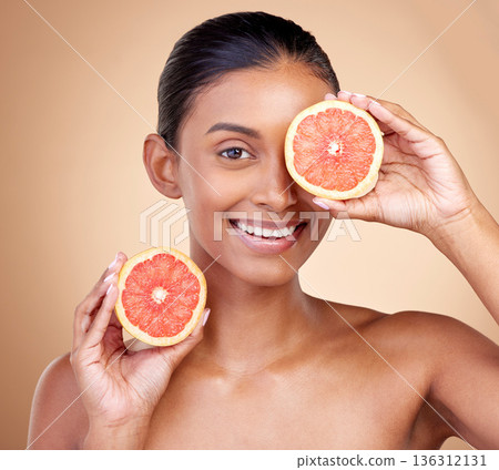 Beauty, woman and grapefruit on eye for skincare, cosmetics and natural product, health or vitamin c in portrait. Face of indian person or model, red fruit and dermatology on studio, brown background Beauty, woman and grapefruit on eye for skincare, cosmetics and natural product, health or vitamin c in portrait. Face of indian person or model, red fruit and dermatology on studio, brown background 136312131