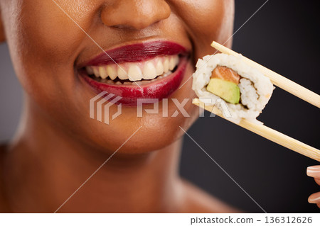 Mouth, eating and woman with sushi and chopsticks in studio for healthy food or teeth. Black female model with makeup on dark background for wellness glow, diet and seafood or salmon closeup Mouth, eating and woman with sushi and chopsticks in studio for healthy food or teeth. Black female model with makeup on dark background for wellness glow, diet and seafood or salmon closeup 136312626