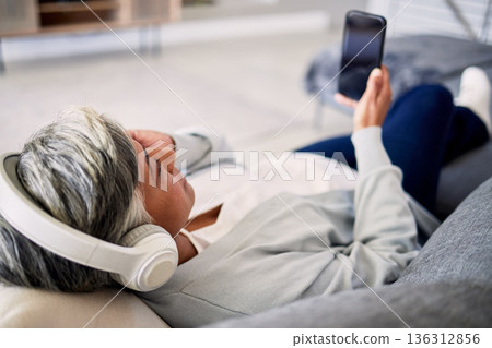 Senior woman, headphones and phone on sofa, relax and music in home living room with search, choice and listening. Elderly lady, smartphone and audio streaming app for radio, podcast and lounge couch Senior woman, headphones and phone on sofa, relax and music in home living room with search, choice and listening. Elderly lady, smartphone and audio streaming app for radio, podcast and lounge couch 136312856