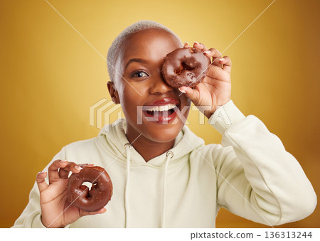 Portrait, chocolate and donut with a black woman in studio on a gold background for candy or unhealthy eating. Smile, food and baking with a happy young female person holding sweet pastry for dessert 136313244
