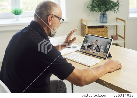 Senior man waving during online videocall on tablet at bright home office Senior man waving during online videocall on tablet at bright home office 136314035