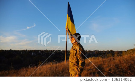Male military in uniform lifting blue-yellow banner in honor of the victory against russian aggression at sunset. Young soldier of ukrainian army raised a waving flag of Ukraine at countryside Male military in uniform lifting blue-yellow banner in honor of the victory against russian aggression at sunset. Young soldier of ukrainian army raised a waving flag of Ukraine at countryside 136314092