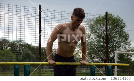 Male shirtless athlete doing push ups on parallel bars at nature. Strong sporty man exercising at sports ground. Young sportsman training hard outdoor. Concept of active sportive lifestyle. Slow mo Male shirtless athlete doing push ups on parallel bars at nature. Strong sporty man exercising at sports ground. Young sportsman training hard outdoor. Concept of active sportive lifestyle. Slow mo 136314103