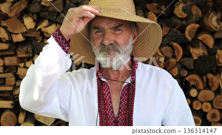 Portrait of senior ukrainian man in straw hat against the background of woodshed. Elderly serious male wearing an embroidered shirt looking into camera outdoor. Cultural heritage and ethnic roots Portrait of senior ukrainian man in straw hat against the background of woodshed. Elderly serious male wearing an embroidered shirt looking into camera outdoor. Cultural heritage and ethnic roots 136314147