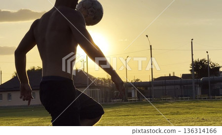 Young man juggling soccer ball on stadium at sunset. Professional footballer kicking ball at green field. Sportsman practicing tricks at meadow with sunlight at background. Freestyle football 136314166