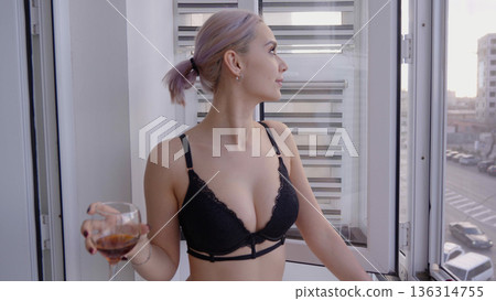 Young woman standing near window with glass of wine 136314755