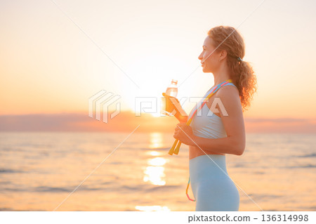 Fit woman drinking water at the beach after running outdoors in the morning. Sporty female relaxing after training. 136314998