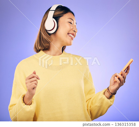 Music, phone and happy Asian woman in studio for social media, internet meme and online chat. Headphones, smile and female person on smartphone listening to audio, song and track on purple background 136315358