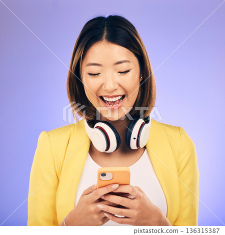 Headphones, phone and happy Asian woman in studio smile for social media, internet meme and chat. Texting, smile and female person on smartphone listen to audio, song and track on purple background 136315387
