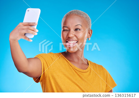 Social media, selfie and funny face with a black woman on blue background in studio to update her profile picture. Post, app and a happy young female influencer taking a photograph for her status Social media, selfie and funny face with a black woman on blue background in studio to update her profile picture. Post, app and a happy young female influencer taking a photograph for her status 136315617