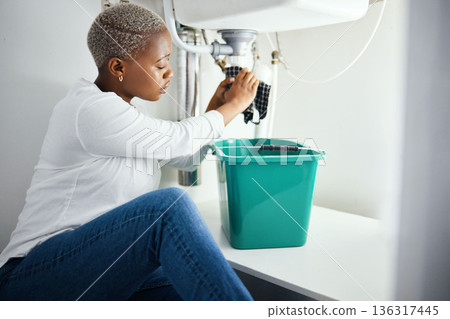 Plumbing, leak and a black woman in the bathroom of her home with a cloth and bucket waiting for assistance. Sink, emergency and a burst pipe with a young female homeowner in her house to stop water 136317445