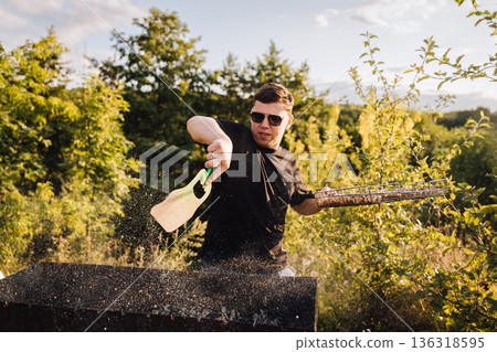 Man fanning charcoal grill outdoors at sunset, sparks flying during summer barbecue 136318595