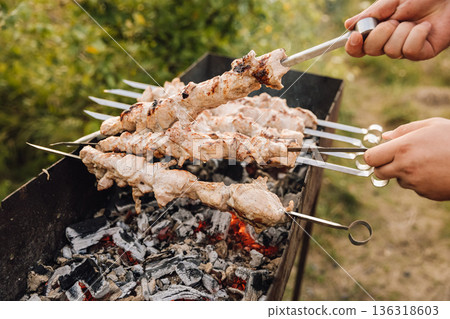 Hands turning pork shashlik skewers over charcoal grill, outdoor barbecue cooking 136318603
