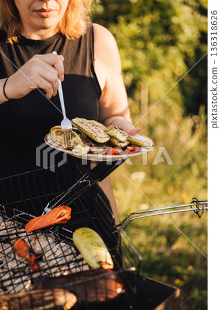 Woman serving grilled vegetables from barbecue, holding plate outdoors in summer sunlight Woman serving grilled vegetables from barbecue, holding plate outdoors in summer sunlight 136318626