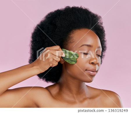 Gua sha, massage and black woman with natural beauty, face care and skin glow on pink background. Female model, jade cosmetic tools and dermatology with afro hair and facial skincare in studio Gua sha, massage and black woman with natural beauty, face care and skin glow on pink background. Female model, jade cosmetic tools and dermatology with afro hair and facial skincare in studio 136319249