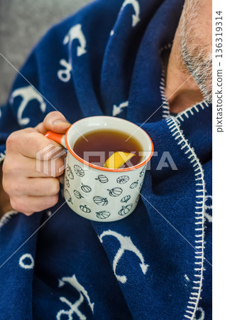 Close-up of a man with a beard wrapped in a soft blue anchor-patterned blanket, holding a warm mug of tea with a slice of lemon.. High quality photo Close-up of a man with a beard wrapped in a soft blue anchor-patterned blanket, holding a warm mug of tea with a slice of lemon.. High quality photo 136319314