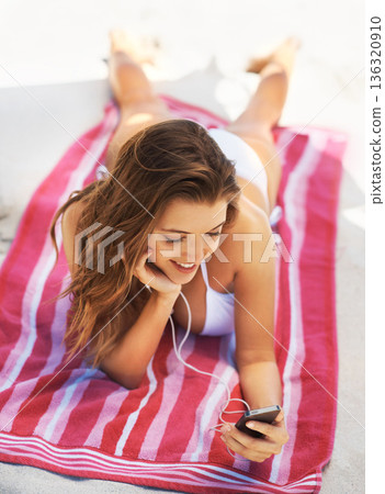 Happy woman, bikini and phone on beach for music in relax for audio listening or sound track outdoors. Female person lying on towel in swimwear enjoying listening to songs on mobile smartphone at sea 136320910