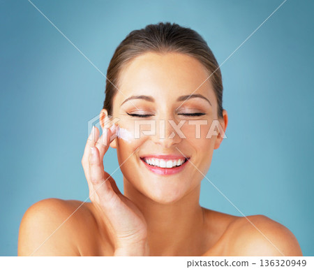 Skincare, smile and woman with cream on face, cosmetics and makeup product on blue background. Happiness, skin care and wellness for happy model with beauty, lotion and dermatology for facial glow. Skincare, smile and woman with cream on face, cosmetics and makeup product on blue background. Happiness, skin care and wellness for happy model with beauty, lotion and dermatology for facial glow. 136320949