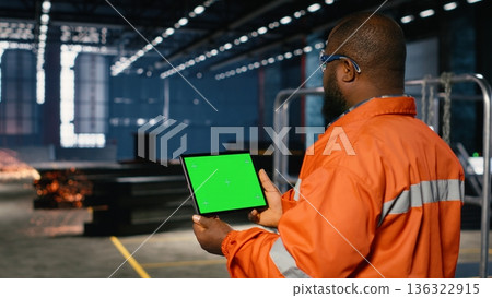 Green screen tablet with worker managing operation tasks across the factory floor, surrounded by metal machinery and fabrication tools that highlight professional engineering. 136322915