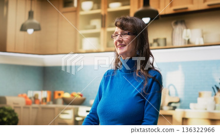 Portrait of joyful aged woman in apartment kitchen shows relaxed enjoyment during retirement. Elder person stands by the counter, embracing casual housekeeping and positive wellbeing. Camera B. 136322966