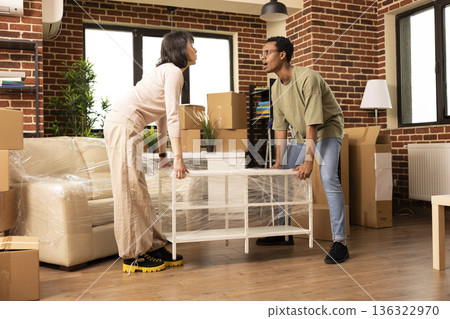Multiethnic couple carries wrapped coffee table into brick wall apartment on moving day. African american husband and caucasian wife arranging furniture and settling into cozy new home. Multiethnic couple carries wrapped coffee table into brick wall apartment on moving day. African american husband and caucasian wife arranging furniture and settling into cozy new home. 136322970