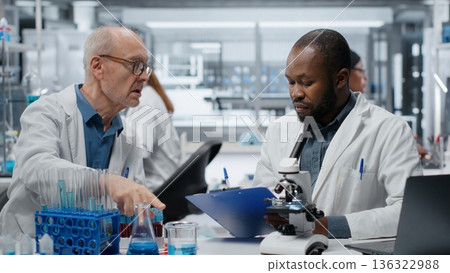 Medical team in lab conducting research on blood sample, taking notes. African american man and elderly colleague in laboratory analyzing sanguine fluid specimen, writing down findings, camera B 136322988