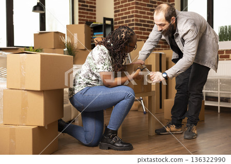 African american woman helping caucasian boyfriend by cutting adhesive tape while packing cardboard boxes. Young multiethnic couple carefully seals cartons, preparing to move out together. African american woman helping caucasian boyfriend by cutting adhesive tape while packing cardboard boxes. Young multiethnic couple carefully seals cartons, preparing to move out together. 136322990