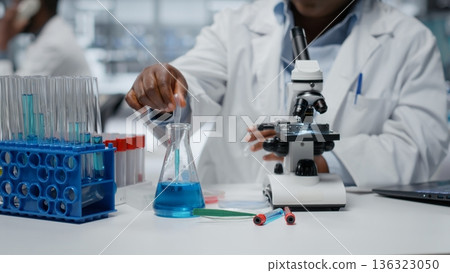 Clinical laboratory technologist pipetting blue liquid into test tube to analyze it. African american woman transferring chemical sample to prepare for immediate spectroscopic analysis, camera B 136323050