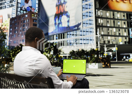 Green screen laptop with black man sitting outdoors on a bench. Person enjoying modern skyscrapers within the metropolitan area of the city center. Trendy street style in a park. Green screen laptop with black man sitting outdoors on a bench. Person enjoying modern skyscrapers within the metropolitan area of the city center. Trendy street style in a park. 136323078