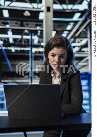Data center female engineer using laptop to review equipment diagnostics. Worker at desk in server room using notebook to adjust configurations, applying firmware updates, cleaning error states 136323086