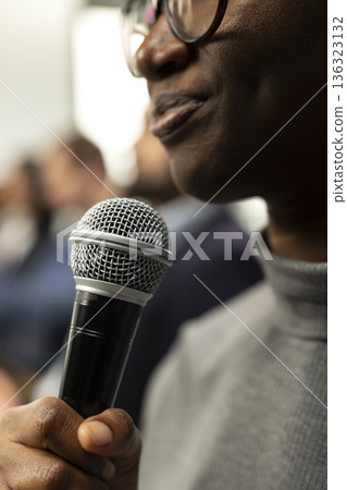 Close up of black woman attendee asking questions on microphone during panel discussion, engaging in debate at international summit. Business conference with presentation on stage. Close up of black woman attendee asking questions on microphone during panel discussion, engaging in debate at international summit. Business conference with presentation on stage. 136323132