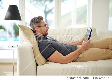 Man, relax and tablet on living room couch, reading and social media for video, game or movies in home. Mature guy, digital touchscreen and lounge sofa for internet blog, news or web app in house 136323386