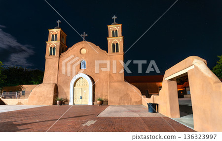 Old San Miguel Mission church in Socorro, New Mexico. Historic adobe architecture features twin towers and a white arch entrance under a starry night sky 136323997