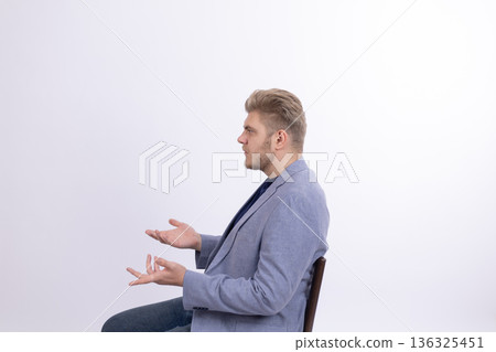 Man in grey suit sitting and gesturing with both hands 136325451