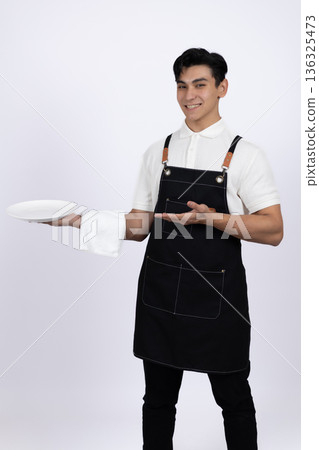 Young male waiter holding a tray and standing in studio 136325473