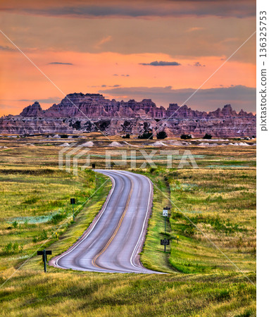 Badlands National Park landscape stands in South Dakota, USA. Scenic view features the winding Loop Road leading towards rugged rock formations and green prairie under a colorful sunset sky Badlands National Park landscape stands in South Dakota, USA. Scenic view features the winding Loop Road leading towards rugged rock formations and green prairie under a colorful sunset sky 136325753