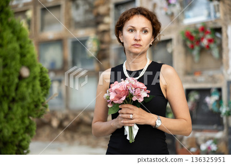 Sorrowful woman in black standing in cemetery with flowers 136325951
